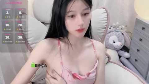 Media: Video of an Asian woman with fair skin, long black hair, and small breasts, wearing a pink bra, sitting on a bed with a stuffed rabbit.