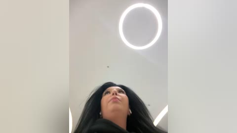 Media: Video of a young woman with long black hair, looking upwards. Her face is partially visible, with a neutral expression. She is indoors under a circular, bright white light fixture. The background is plain and white.