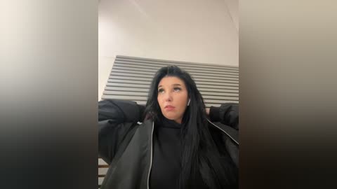 Media: Video of a young woman with long black hair, wearing a black leather jacket, standing against a white wall with horizontal blinds.