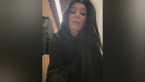 Media: Video of a woman with long black hair, wearing a dark jacket, standing in a narrow hallway with beige walls and a ceiling light. Her expression is neutral.