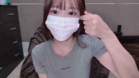 Media: A video of an Asian woman with long brown hair and a white surgical mask, wearing a grey t-shirt, seated indoors. She adjusts her mask with her right hand.