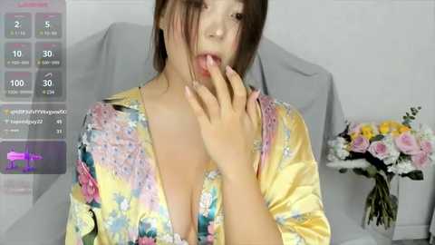 Media: Video of an Asian woman with light skin, brown hair, and a low-cut floral blouse, licking her fingers in a suggestive manner. Background shows a gray cushion and a bouquet of flowers.