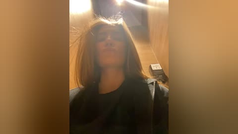 Media: Video of a young woman with long brown hair, wearing a black jacket, standing in a dimly lit, wooden-paneled room with a TV and phone visible in the background.