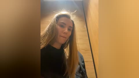 Media: Video of a young Caucasian woman with long blonde hair, wearing a black top, standing in a narrow, wooden-paneled hallway. The lighting is warm, creating a cozy atmosphere.