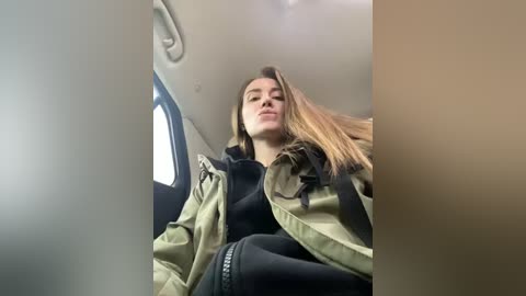 Media: Video of a young woman with light skin, long brown hair, and a slim physique, wearing a green jacket and black pants, seated inside a car. The background shows the car's interior.