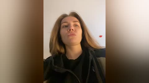 Media: Video of a Caucasian woman with straight, light brown hair, wearing a black jacket, making a kissy face. The background is a blurry, white wall with a red object partially visible.