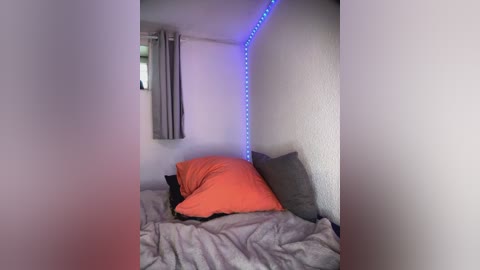 Media: A video of a small, dimly lit room with a single bed covered in gray sheets, featuring an orange pillow and a gray pillow, and a blue LED strip lighting the wall.