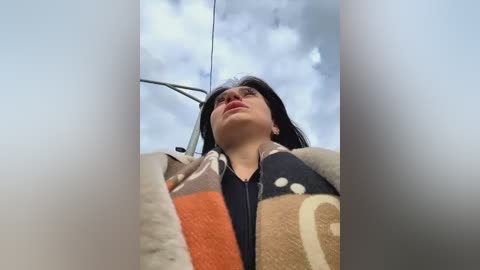 Media: Video of a woman with long black hair, wearing a beige coat and scarf with orange and white patterns, looking up at a cloudy sky.