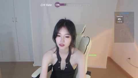 Media: Video of an Asian woman with fair skin and black hair, wearing a black halter top, sitting in a modern white chair. Background includes white cabinets and a green-lit gaming setup.