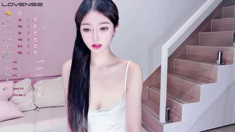 Media: Video of an Asian woman with long black hair, fair skin, and red lipstick, wearing a white lace camisole, standing in a modern, minimalist living room with light pink walls and beige sofa.