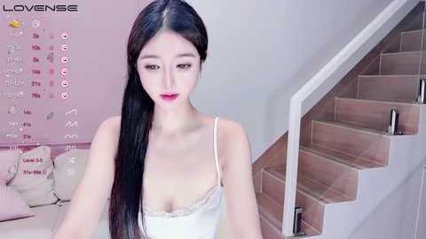 Media: Video of a young East Asian woman with long black hair, fair skin, and pink lipstick, wearing a white lace camisole, sitting on a pink sofa in a modern, pastel-toned living room.