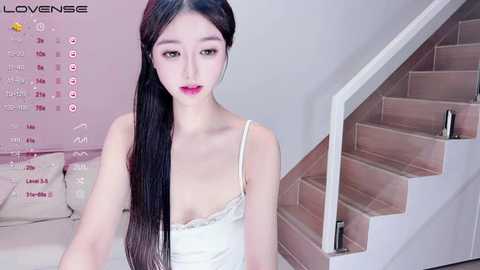 Media: Video of a young East Asian woman with long, straight black hair, fair skin, and small breasts, wearing a white lace camisole, sitting on a staircase in a modern, light-toned room.