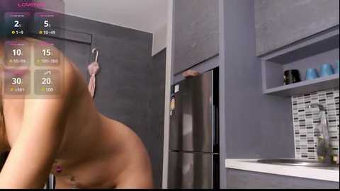 Media: Video of a nude person with a tan body, bending over, in a modern kitchen with gray cabinets, white counters, and a stainless steel fridge.