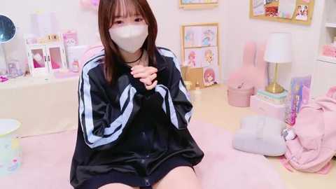 Media: A video of a young Asian woman with long brown hair and a white face mask, sitting in a brightly lit, pink-themed room with pastel-colored toys and furniture.