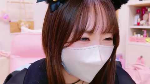 Media: Video of an Asian woman with straight brown hair, wearing black cat ears, a white face mask, and a black robe, standing in a pink-themed room with shelves and storage boxes in the background.