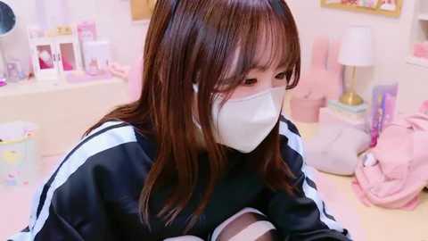 Media: Video of a young Asian woman with long brown hair and bangs, wearing a white face mask, black jacket, and sitting at a desk in a soft, pastel-toned room with pink and white decor.