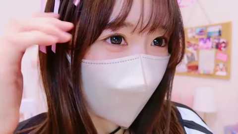 Media: Video of an Asian woman with long brown hair and light skin, wearing a white face mask, pink headphones, and a black and white striped shirt, in a bright, cluttered room with a bulletin board and pastel-colored walls.