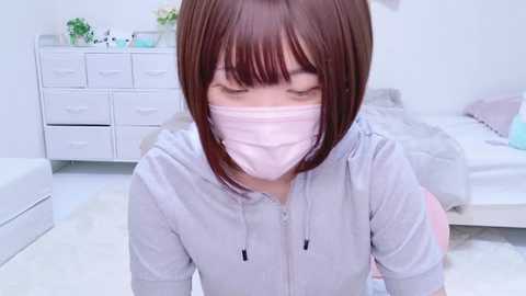 Media: Video of an East Asian woman with straight brown hair and bangs, wearing a pink face mask, light gray hoodie, and white shirt, in a white room with a bed, dresser, and potted plant.