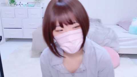 Media: Video of an East Asian woman with short brown hair, wearing a pink surgical mask, sitting on a light-colored rug in a minimalist, white bedroom.