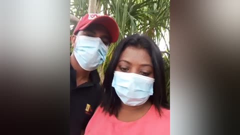 Media: Video of two people wearing light blue medical masks outdoors, surrounded by lush green palm leaves. One wears a red cap, and the other a pink shirt, suggesting a casual, protective setting.