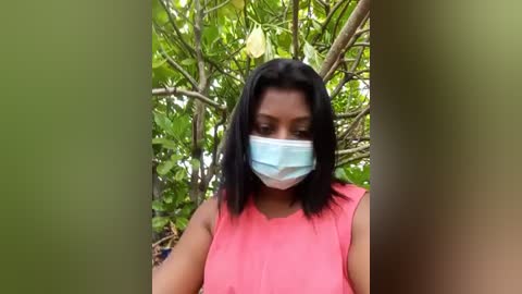 Media: Video of a young woman with medium skin tone and shoulder-length black hair, wearing a pink sleeveless top and a light blue surgical mask, standing in a lush, green outdoor setting with dense foliage and a yellow flower in the background.