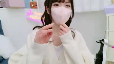 Media: Video of a young East Asian woman with long brown hair, wearing a white face mask, a cream sweater, and a white top, adjusting her mask in a softly lit, minimalist bedroom with pastel accents and a pumpkin decoration.