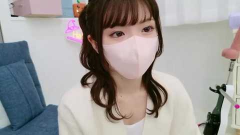 Media: Video of an Asian woman with long brown hair, wearing a white mask and robe, seated in a brightly lit room with a blue chair and pink wall decorations.