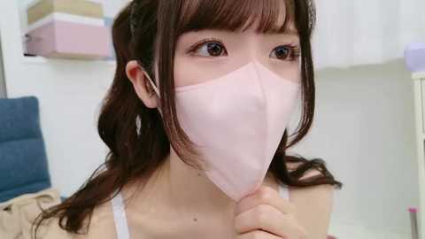 Media: A video of an Asian woman with long, dark hair in pigtails, wearing a pink surgical mask, light-colored top, and a blue chair in the background.