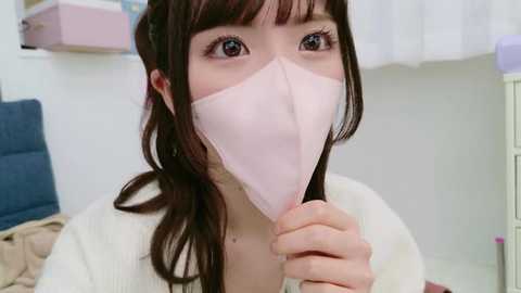 Media: Video of an Asian woman with light skin, long dark brown hair, and bangs, wearing a light pink face mask, holding it, in a white shirt, against a blurred indoor background.