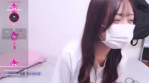 Media: Video of an East Asian woman with long dark hair and a white face mask, wearing a white lab coat, sitting on a white bed, looking distressed, in a clinical setting.