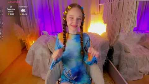 Media: Video of a young girl with light skin, twin braids, and a blue tie-dye top, smiling in a cozy, dimly-lit room with purple and yellow lights, macrame decor, and a gray couch.