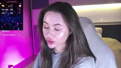 Media: Video of a young woman with long brown hair, wearing a light gray sweater, sitting on a white chair, in a modern bedroom with purple lighting and a bed in the background.