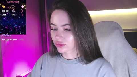 Media: Video of a young woman with long dark hair, wearing a light gray sweater, seated indoors with a purple and pink ambient light. Background includes a digital display showing \"Landon\" with weather and time details.