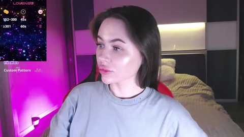 Media: Video of a young woman with straight black hair, fair skin, and light blue shirt, sitting in a dimly lit room with purple and pink lighting. Behind her, a bed with a striped blanket.