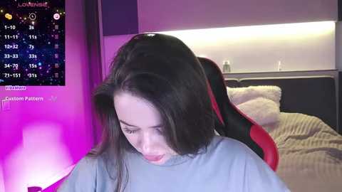 Media: Video of a young woman with long black hair, wearing a gray shirt, sitting in a gaming chair in a dimly lit room with purple lighting.