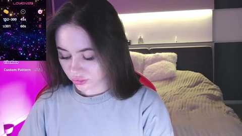 Media: Video of a young woman with straight black hair, wearing a gray sweater, sitting on a bed with a beige blanket and a stuffed animal. Background shows a night sky on a digital device.