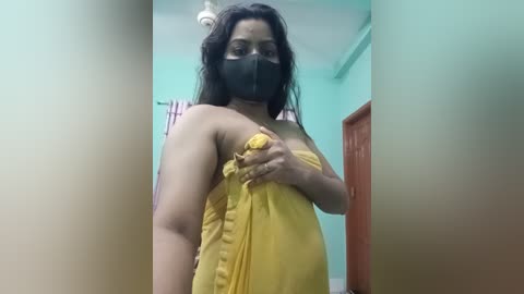 Media: Video of a pregnant woman with medium brown skin, long black hair, wearing a yellow strapless dress, black face mask, and holding a small stuffed toy. She stands in a bathroom with teal walls, a towel rack, and a closed door.