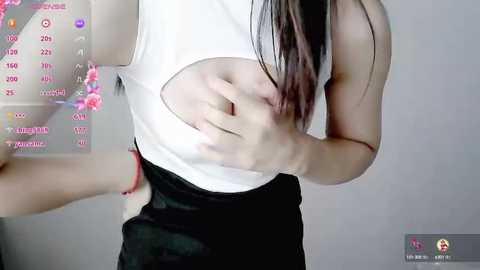 Media: Video of a light-skinned woman with long dark hair, wearing a white tank top and black skirt, holding her stomach. Background includes a pink drink and a gray wall.