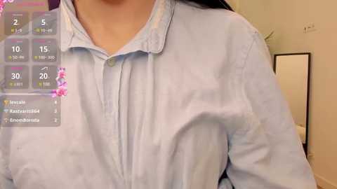 Media: Video of a woman's upper torso wearing a light blue button-up shirt, with a virtual AR overlay showing menstrual cycle tracking. Background shows a simple room with a white wall and a mirror.