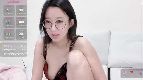 Media: Video of an East Asian woman with fair skin, long black hair, wearing glasses, and a black lace bra, sitting on a white couch, with a virtual reality game display in the background.