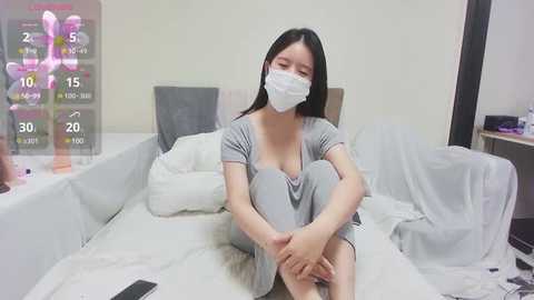 Media: Video of an East Asian woman with long black hair, wearing a white mask and gray pajamas, sitting on a white bed, surrounded by medical devices and a TV displaying vital signs.