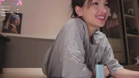 Media: Video of a young woman with medium skin tone, wearing a grey robe, smiling, holding a blue cup, sitting on a wooden floor, with a TV showing a man in the background.