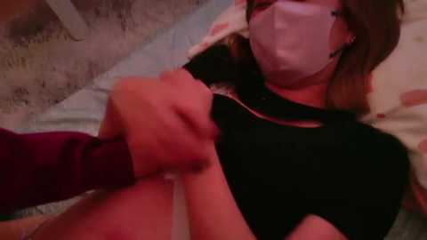 Media: A video of a woman with light skin, brown hair, and a white mask lying on a bed, being held down by a person's arm. She is wearing a black shirt.