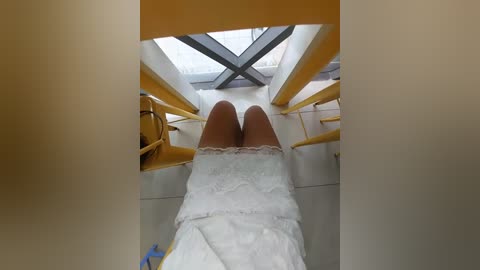 Media: Video of a person's legs wearing white lace-trimmed socks and a white dress, lying on a yellow chair with geometric metal supports, in a modern, minimalist room with large windows.