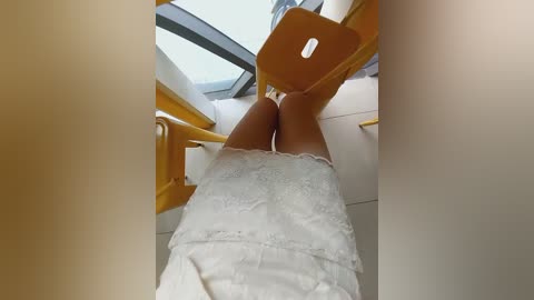 Media: A video of a woman's legs in a white lace dress, seated on a yellow chair, with a glass window and white walls in the background.