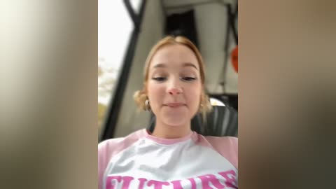 Media: Video of a young woman with light skin and blonde hair, wearing a pink and white T-shirt with \"FUTURE\" in pink text, sitting in a car. The background shows a blurred outdoor scene.