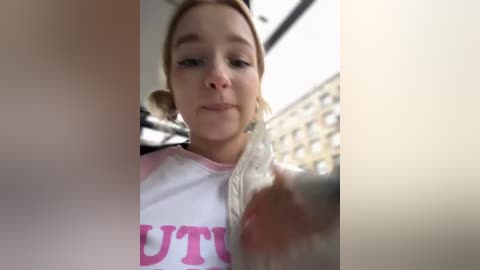 Media: A video of a young woman with light skin, blonde hair, and light makeup, wearing a white T-shirt with pink text, sitting in a car. The background shows an out-of-focus cityscape.