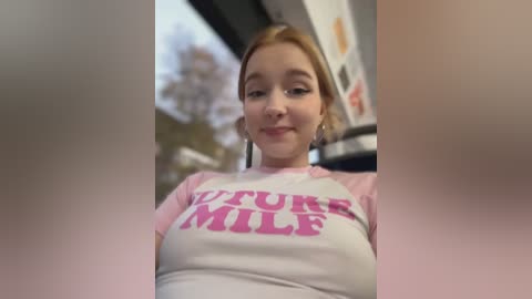 Media: Video of a young Caucasian woman with light skin and blonde hair, wearing a pink and white long-sleeve shirt with \"NEVER TOO OLD TO MILK\" printed in pink. She's smiling. Background shows blurred outdoor setting.