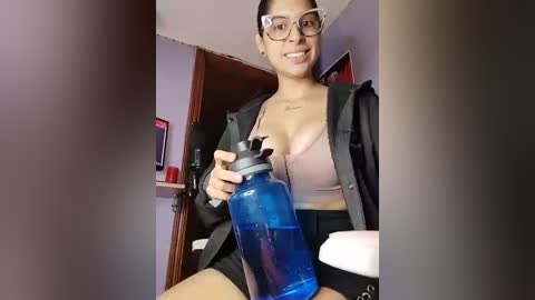 Media: Video of a smiling Latina woman with glasses and a small tattoo on her neck, wearing a sports bra and black jacket, holding a blue water bottle in a dimly lit room.