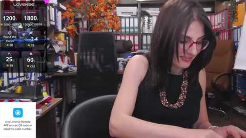 Media: Video of a woman with shoulder-length dark hair, glasses, wearing a black top, seated at a cluttered desk in a busy office.